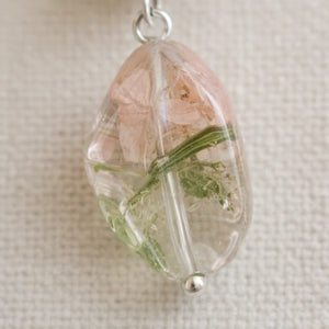 Close‑up of a clear resin pendant containing dried pink petals and green botanical elements, attached to a small metal loop and displayed on textured beige fabric.