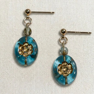 Handmade earrings featuring a faceted gray bead and an oval blue glass bead with a gold floral motif, displayed on light fabric.