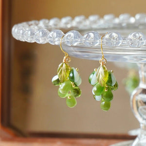 Close-up of green glass grape cluster dangle earrings with gold leaves hanging on a glass dish rim