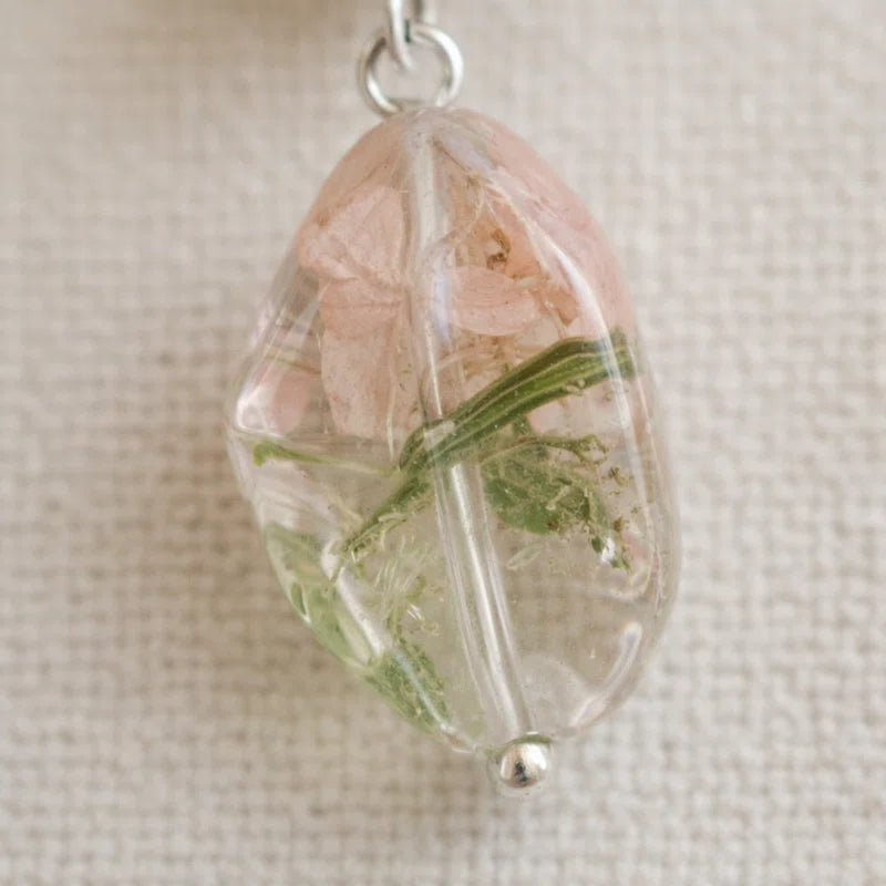 Close‑up of a clear resin pendant containing dried pink petals and green botanical elements, attached to a small metal loop and displayed on textured beige fabric.
