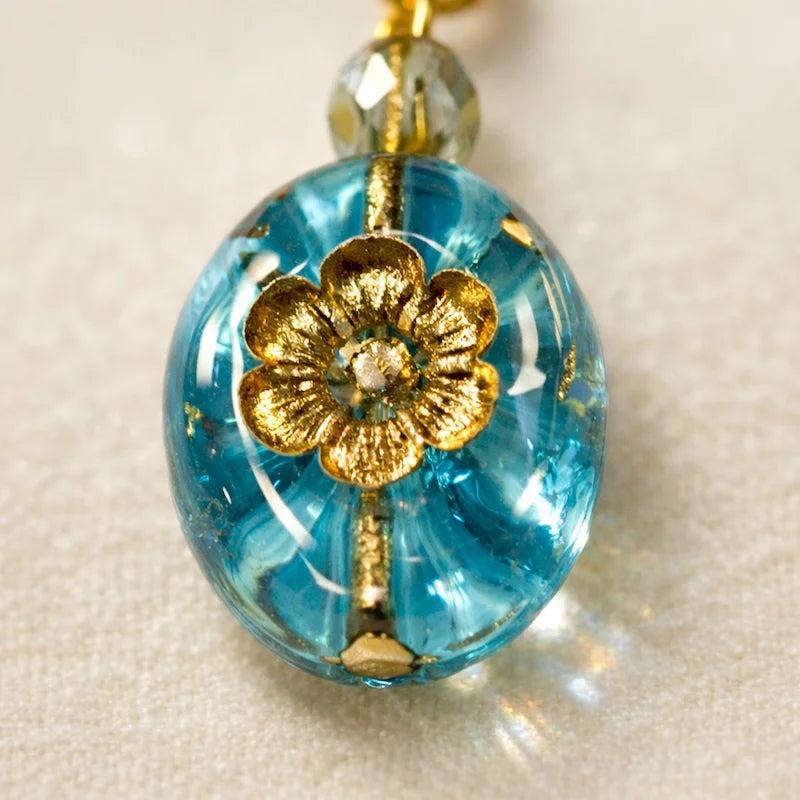 Close‑up of a handmade blue glass pendant featuring a gold floral motif and a faceted metallic bead, displayed on soft light‑colored fabric.