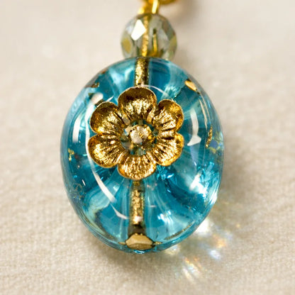Close‑up of a handmade blue glass pendant featuring a gold floral motif and a faceted metallic bead, displayed on soft light‑colored fabric.