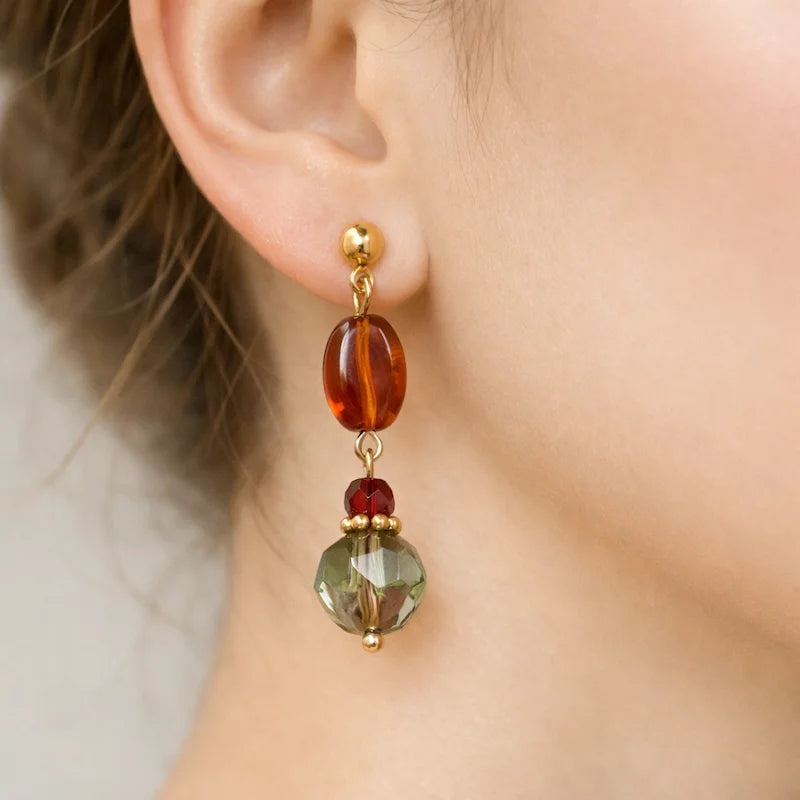 Close‑up of an ear wearing a handmade earring featuring a gold stud, an amber oval bead, a small red bead, and a larger faceted green bead, connected with gold accents against a soft neutral background.