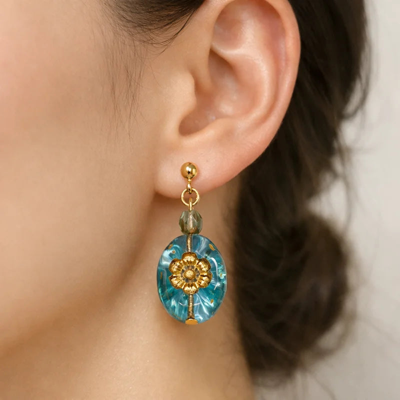 Close‑up of an ear wearing a handmade earring featuring a translucent blue oval pendant with a gold floral motif and a small faceted bead, connected by gold metal hardware against a soft neutral background.