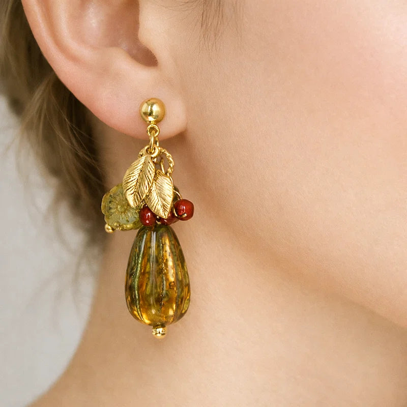 Close‑up of an ear wearing a handmade earring featuring gold leaf charms, red accent beads, a textured yellow‑green bead, and a large amber marbled teardrop pendant, shown against a soft neutral background.