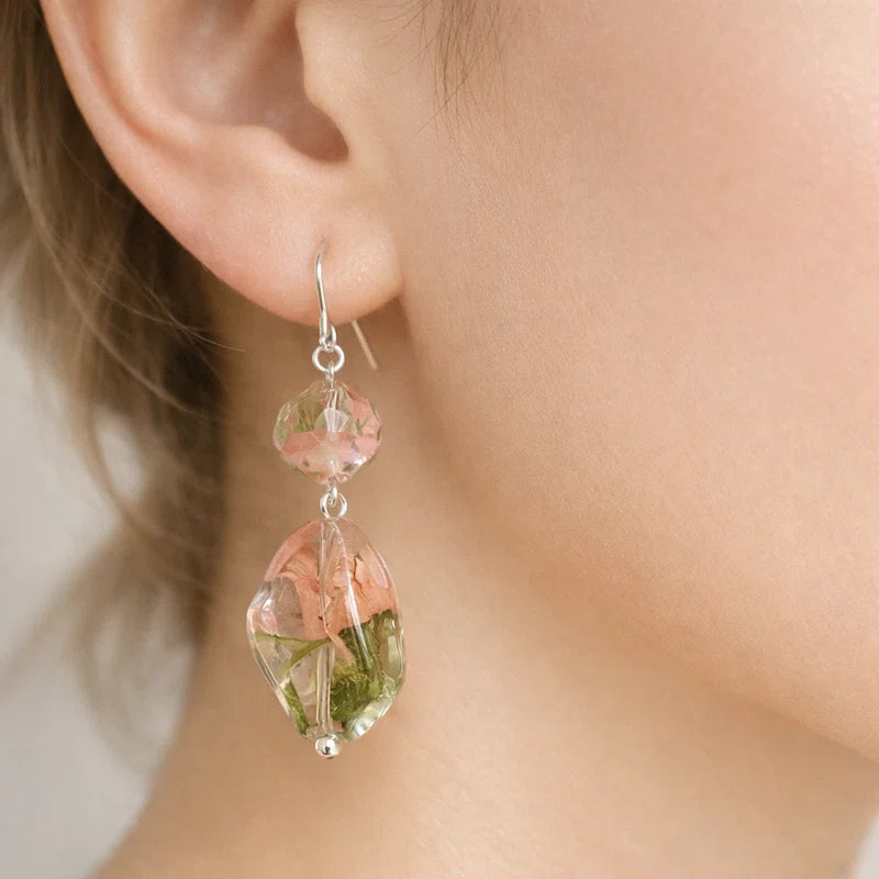 Close‑up of an ear wearing a handmade earring featuring two clear resin beads with embedded dried pink and green flowers, connected with silver hardware against a soft neutral background.