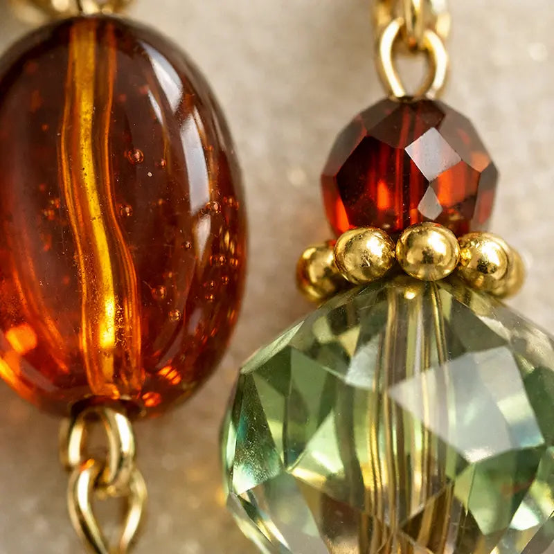 Close‑up of two connected beads: an amber oval glass bead with internal bubbles and a gold stripe, paired with a faceted red bead and a larger faceted green bead separated by gold accents, displayed on a soft neutral surface.