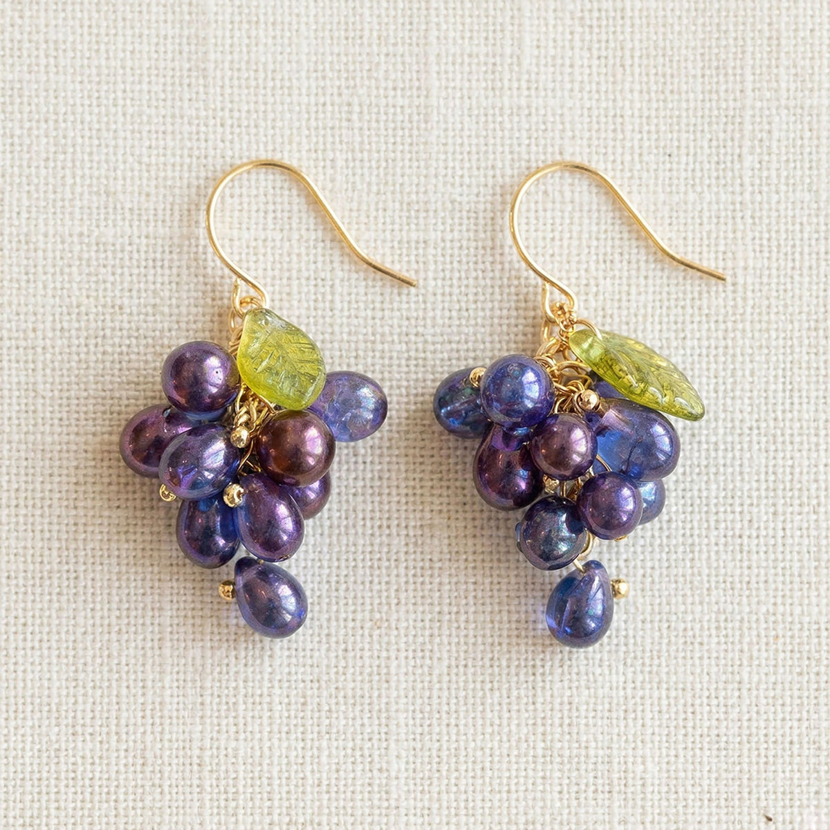 Flatlay of purple grape cluster earrings on unbleached linen fabric with antique bronze hooks