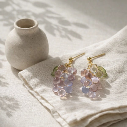 Grape‑cluster earrings made of translucent purple and blue beads with green leaf accents, displayed on folded beige fabric beside a matte ceramic vase in soft natural lighting.