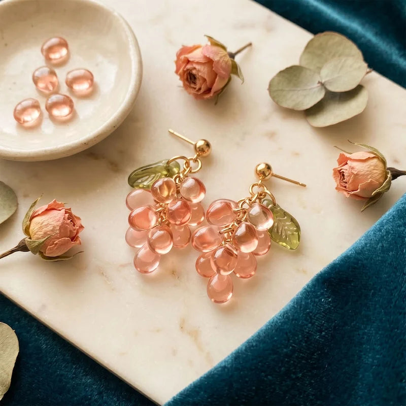 Handmade Czech glass earrings with translucent pink grape beads and green leaf accents displayed with dried roses, eucalyptus and velvet fabric, romantic nature‑inspired jewelry for women