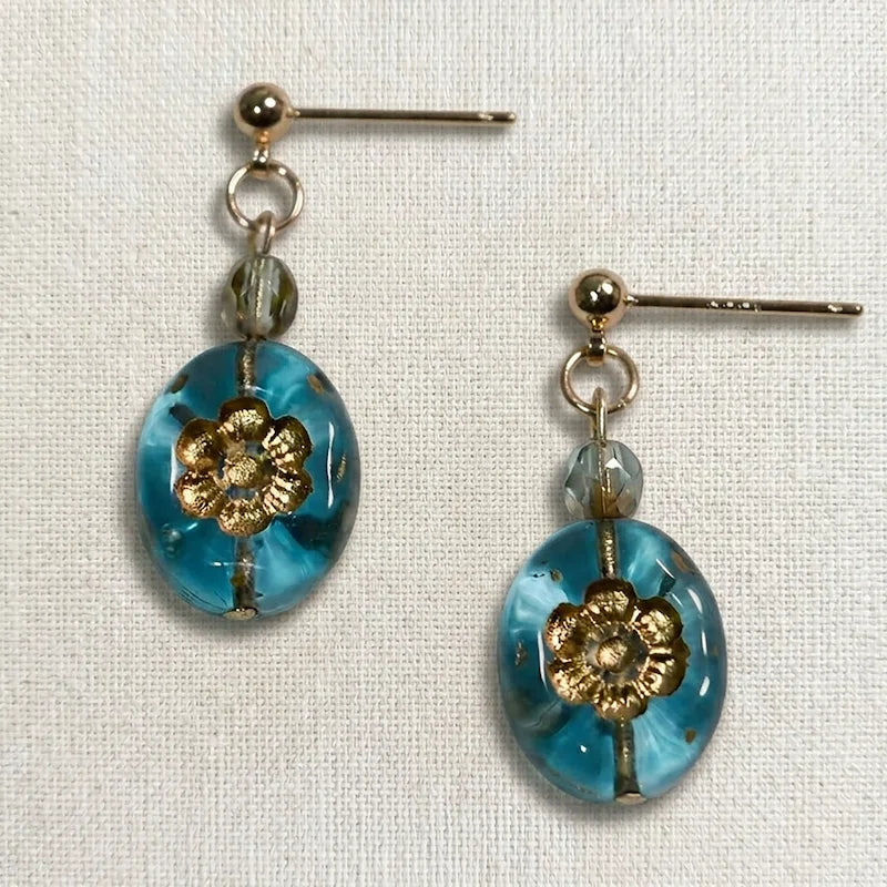 Handmade earrings featuring a faceted gray bead and an oval blue glass bead with a gold floral motif, displayed on light fabric.