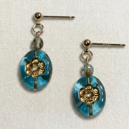 Handmade earrings featuring a faceted gray bead and an oval blue glass bead with a gold floral motif, displayed on light fabric.