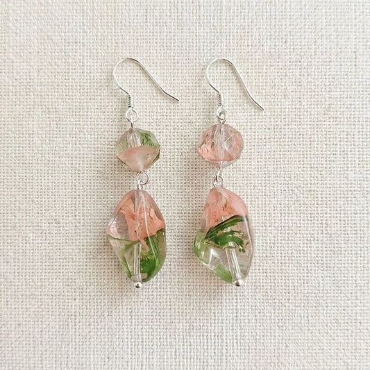 Handmade earrings featuring a faceted pink‑green resin bead and a larger clear resin drop with embedded dried flowers, displayed on light beige textured fabric.