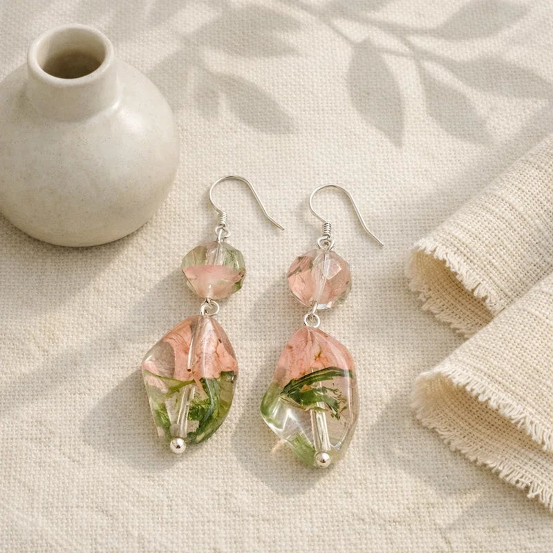 Handmade earrings featuring faceted pink‑green resin beads and larger clear resin drops with embedded dried flowers, displayed on beige fabric beside a small ceramic vase under soft natural light.