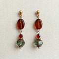 Handmade earrings featuring gold ball studs, oval amber beads, faceted red beads, and larger green‑toned faceted beads, arranged symmetrically and displayed on light fabric.