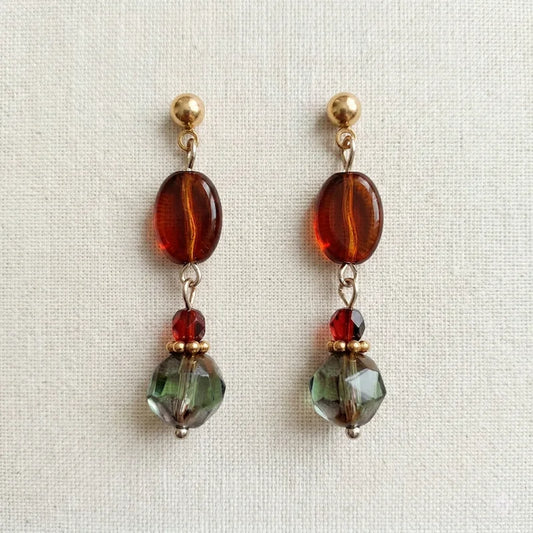 Handmade earrings featuring gold ball studs, oval amber beads, faceted red beads, and larger green‑toned faceted beads, arranged symmetrically and displayed on light fabric.