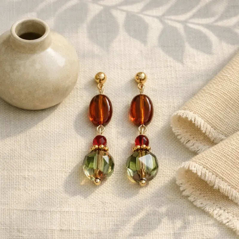 Handmade earrings featuring gold studs, amber oval beads, small red beads, and large faceted green beads, displayed on beige textured fabric beside a small ceramic vase and folded textiles under soft natural light.