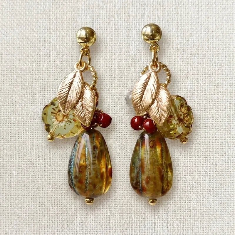 Handmade earrings featuring gold studs, metallic leaf charms, translucent yellow‑green floral beads, red accent beads, and large marbled teardrop beads in amber, brown, and blue tones, displayed on light fabric.