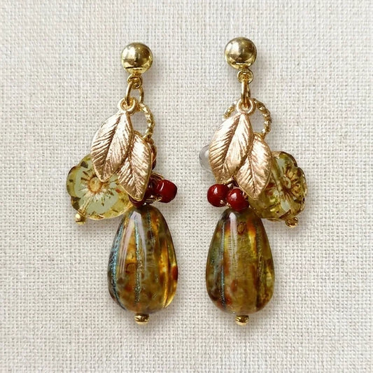 Handmade earrings featuring gold studs, metallic leaf charms, translucent yellow‑green floral beads, red accent beads, and large marbled teardrop beads in amber, brown, and blue tones, displayed on light fabric.