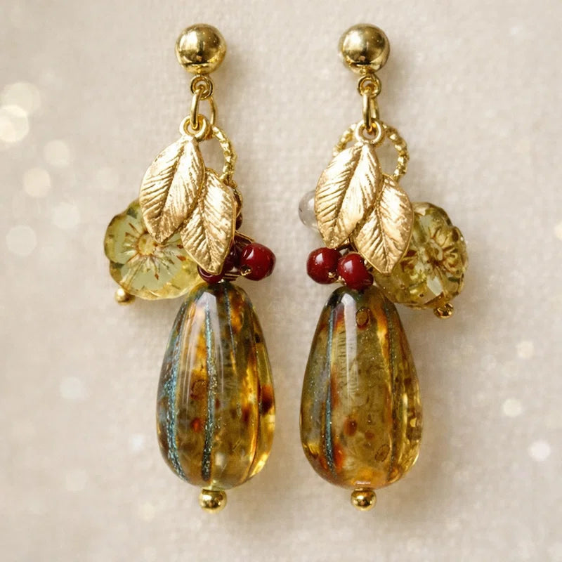 Handmade earrings featuring gold studs, small gold leaf charms, translucent light‑green floral beads, red accent beads, and large amber‑toned marbled teardrop beads, displayed on light fabric.