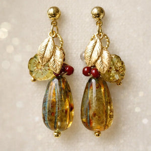 Handmade earrings featuring gold studs, small gold leaf charms, translucent light‑green floral beads, red accent beads, and large amber‑toned marbled teardrop beads, displayed on light fabric.