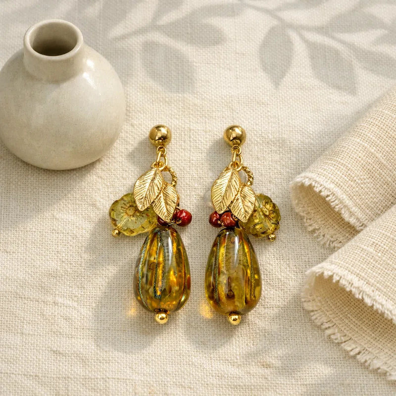 Handmade earrings featuring gold studs, small metallic leaf charms, translucent yellow‑green beads, red accent beads, and large amber marbled teardrop beads, displayed on beige fabric beside a small ceramic vase and rolled textiles.