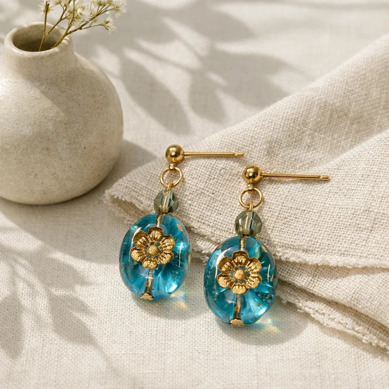 Handmade earrings featuring translucent green beads and oval blue resin pieces with embedded gold floral designs, displayed on beige fabric beside a small ceramic vase with dried flowers.