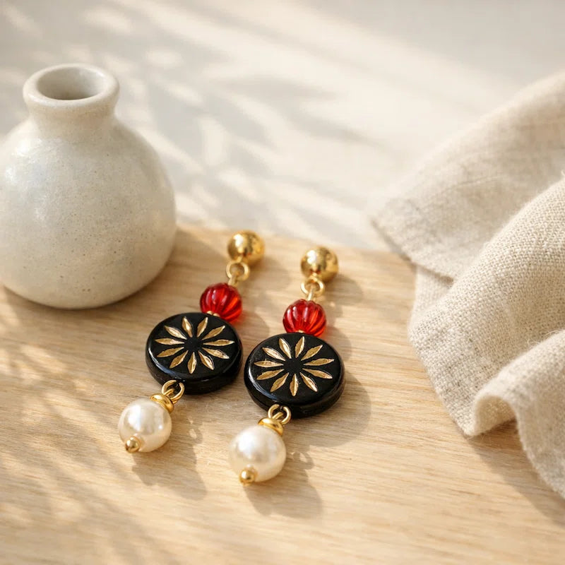 Handmade earrings with red bead, black gold floral medallion and white pearl drop displayed on wooden surface, elegant jewelry for women