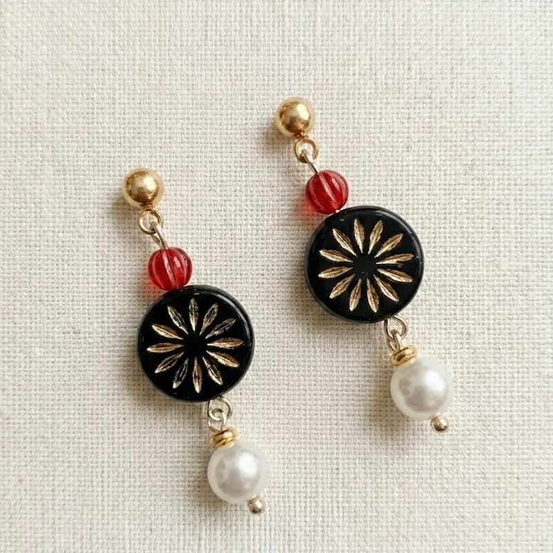 “Handmade earrings with red bead, black gold floral medallion and white pearl drop on fabric background, elegant jewelry for women