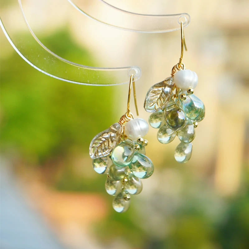 Close-up of green glass grape cluster dangle earrings with pearl beads and gold leaf charms, hanging from a clear acrylic stand