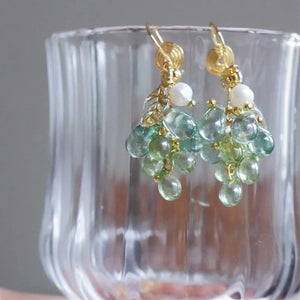 Green glass grape cluster dangle earrings with white pearls and gold leaf charms, draped over the rim of a clear ribbed glass vase