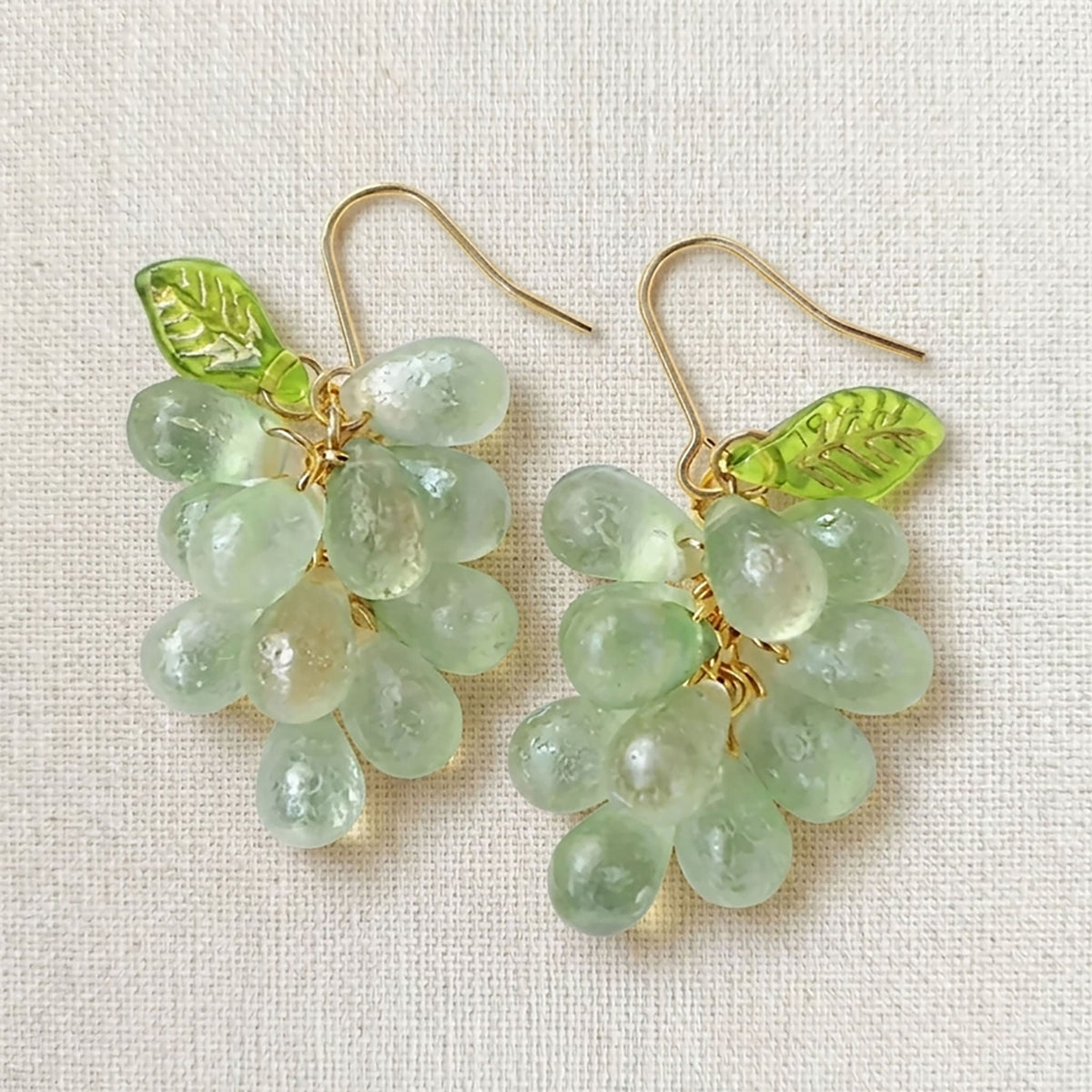 Pair of handmade purple glass grape drop earrings with gold hooks on a linen background